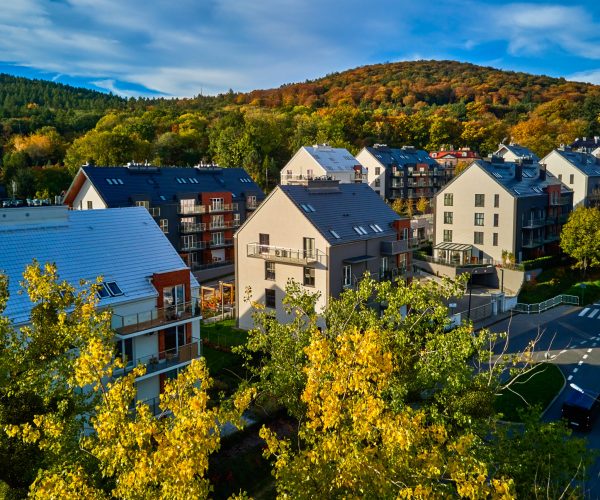 Residential building facade with balconies and windows near vibrant autumn forest, aerial view. Contemporary apartments in suburban eco friendly neighborhood with green spaces near mountains